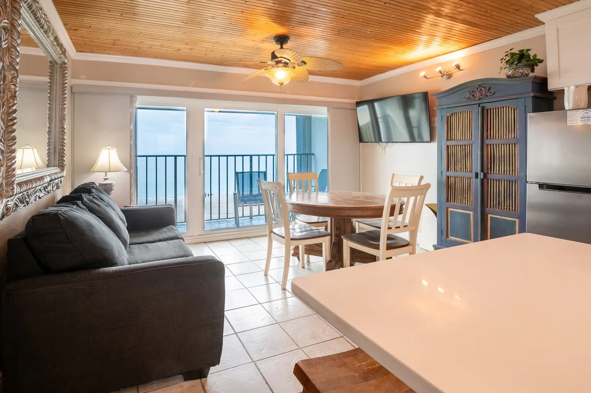 A sunlit living room with a comfortable couch and large windows framing panoramic ocean views. The round dining table for four sits near the window, and coastal-inspired décor—such as beadboard ceilings and a decorative cabinet—enhance the inviting, beachy atmosphere.