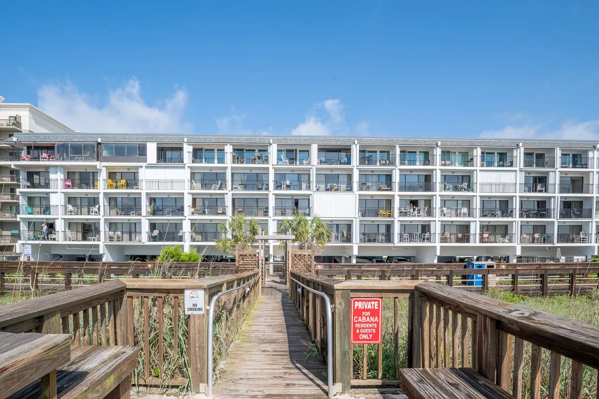 An exterior perspective showcasing a welcoming boardwalk leading directly to the building, with the facade and cheerful balconies visible. The scene hints at easy access to both indoor comforts and beach activities.