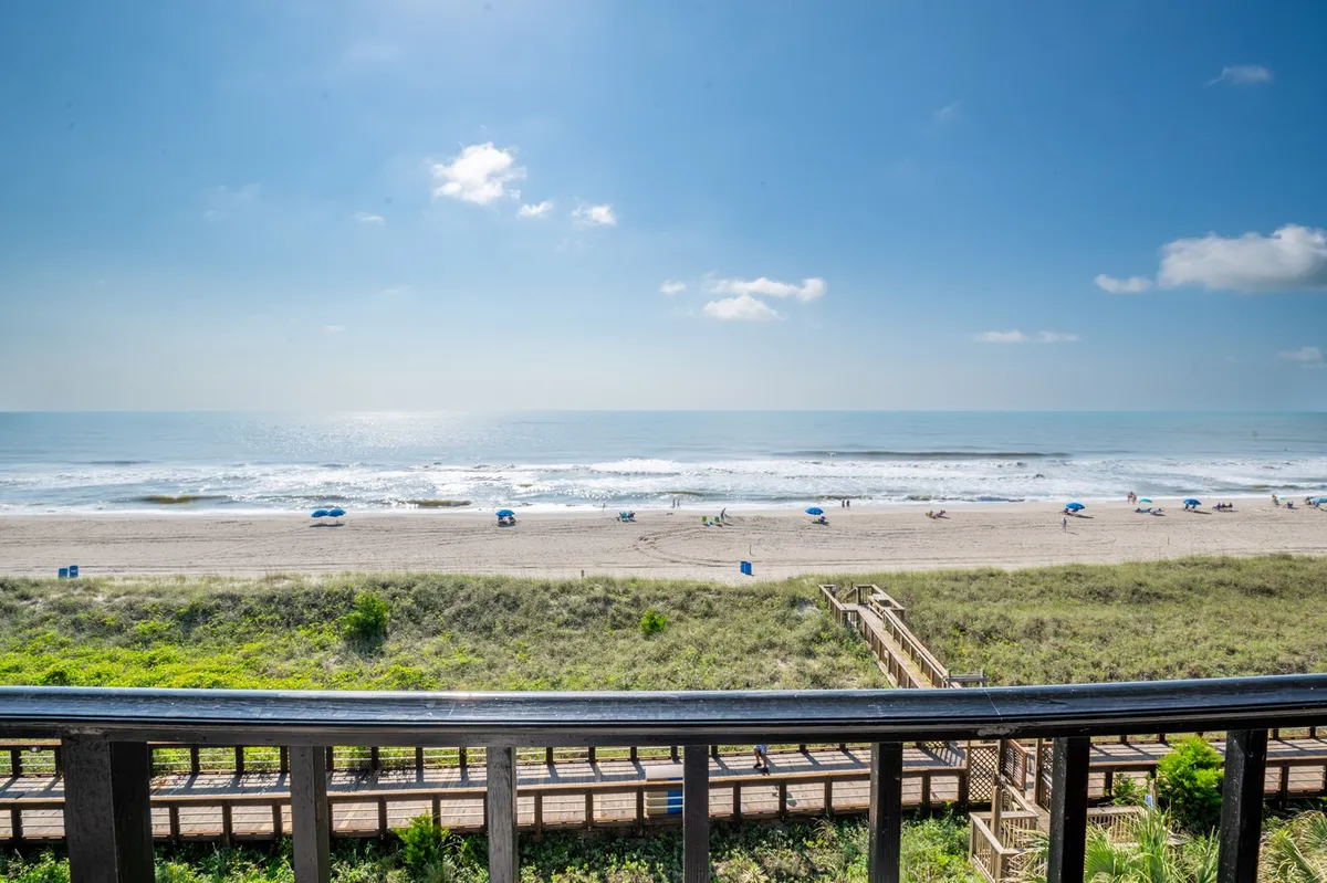 A vibrant view of the shoreline with gentle ocean waves, dotted with colorful beach umbrellas and a wooden boardwalk running parallel to the sand. The scene is lively with families sunbathing, swimming, and enjoying picnics, capturing the joyful spirit of a classic beachside community.