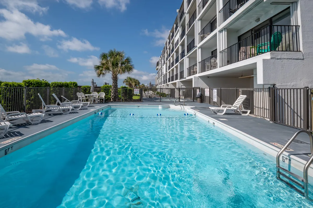 A vibrant exterior view of the sparkling swimming pool surrounded by comfortable poolside loungers and lush palm trees. The condo balconies overlook the pool area, adding to the inviting, resort-like ambiance.