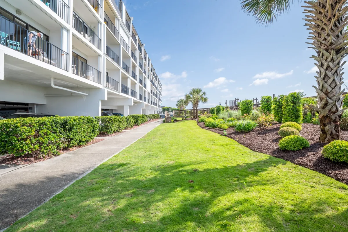 A vibrant exterior scene featuring a lush green lawn, palm trees, colorful flower beds, and multiple balconies overlooking the landscape. Families and groups are seen strolling, relaxing, and gathering outdoors, suggesting a tranquil, communal setting just steps from the ocean.