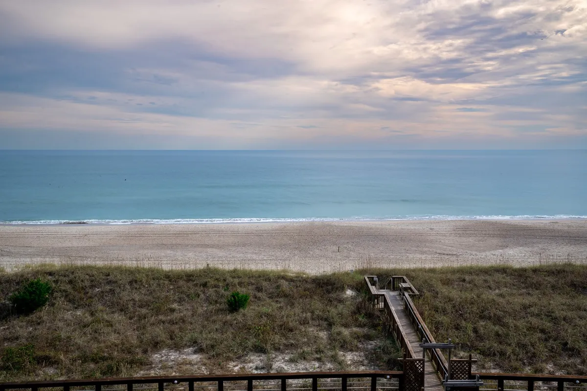 A serene view of a sandy beach and calm sea, with a wooden walkway leading towards the horizon. The scene invites visitors to stroll along the shore, watch the sunset, and enjoy the soothing sounds of waves, perfectly encapsulating the natural beauty accessible from the condo.