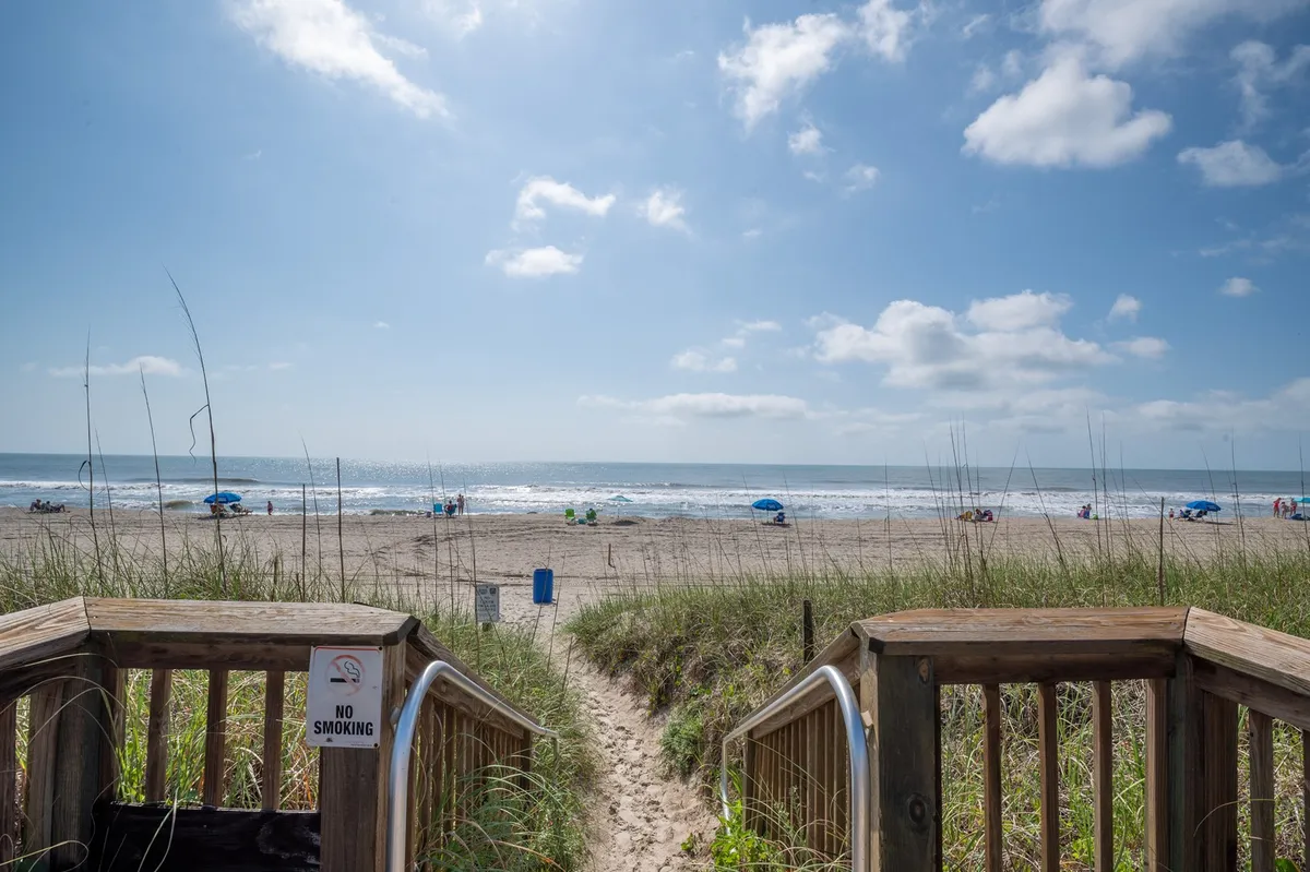 A sweeping view of the wooden stairs leading directly to a wide sandy beach and the ocean beyond. The sky is filled with soft clouds, and the shoreline invites families to play, sunbathe, and enjoy sandcastle building. The perspective showcases easy, direct access to the beach and the beauty of the coastal environment—key elements for family beach days.
