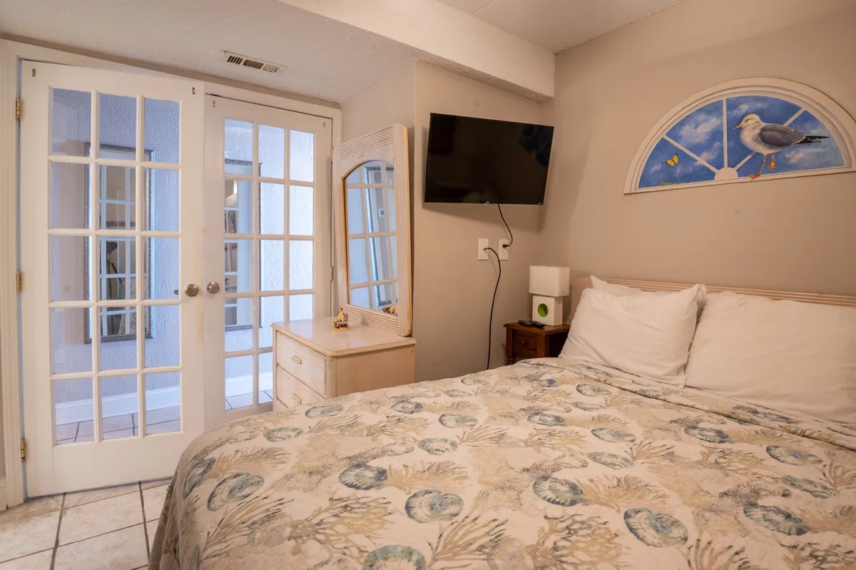 This image depicts a stylish primary bedroom featuring a plush queen bed with patterned bedding, a wall-mounted smart TV, French doors leading to an outdoor space, and a decorative mirror. The natural light streaming in and the warm, inviting decor create a relaxing atmosphere.