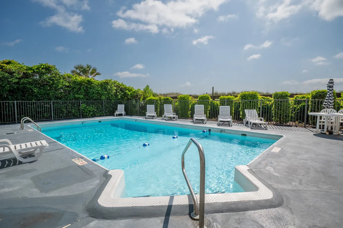 A resort-style swimming pool surrounded by lounge chairs and lush greenery, under open skies. The pool area is spacious and inviting, with plenty of room for families to relax, swim, and play together during sunny afternoons. The scene suggests a leisurely atmosphere perfect for cooling off after a beach day.
