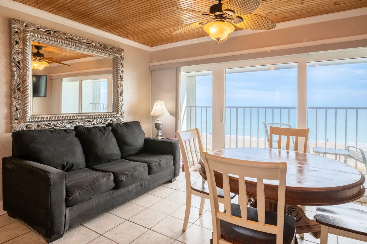 The image showcases the condo's inviting open-concept living and dining area, featuring a comfortable sofa and a dining table positioned to maximize the breathtaking ocean view visible through expansive floor-to-ceiling windows. A large mirror amplifies the coastal light, making the space feel bright and airy, while the layout encourages relaxation, conversation, and family meals with the sea as your backdrop.