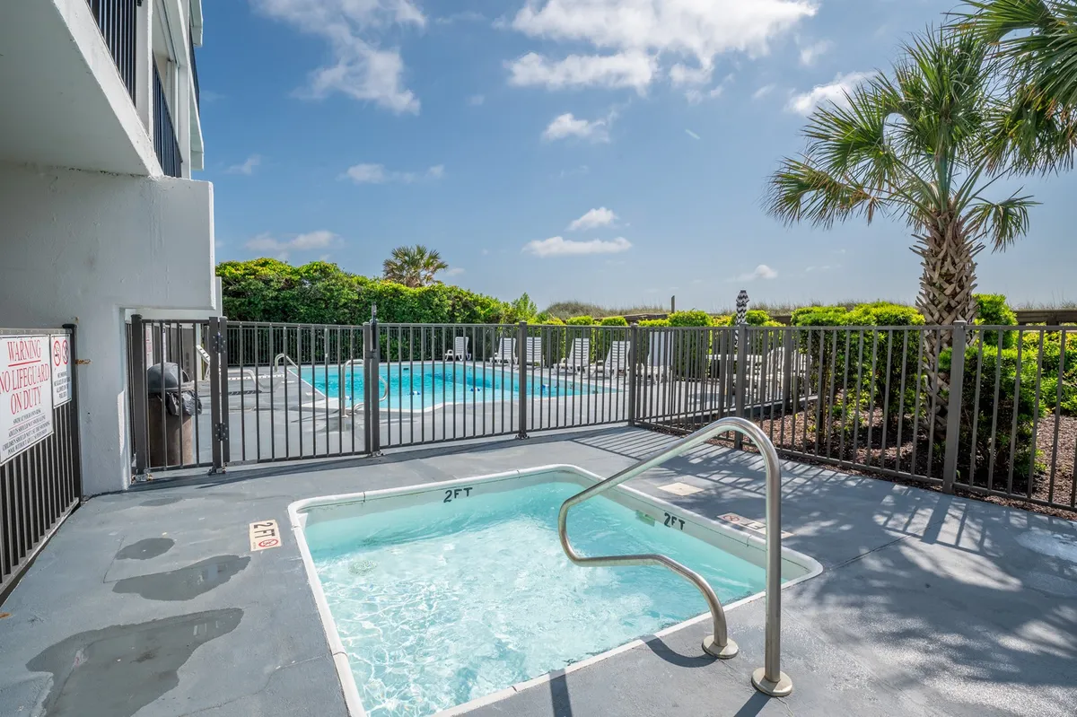 A lively pool area featuring a relaxing hot tub, swimming pool, and lush palm trees, all enclosed by a safety fence. The inviting atmosphere encourages group gatherings, swimming, and tranquil moments under the evening sky.