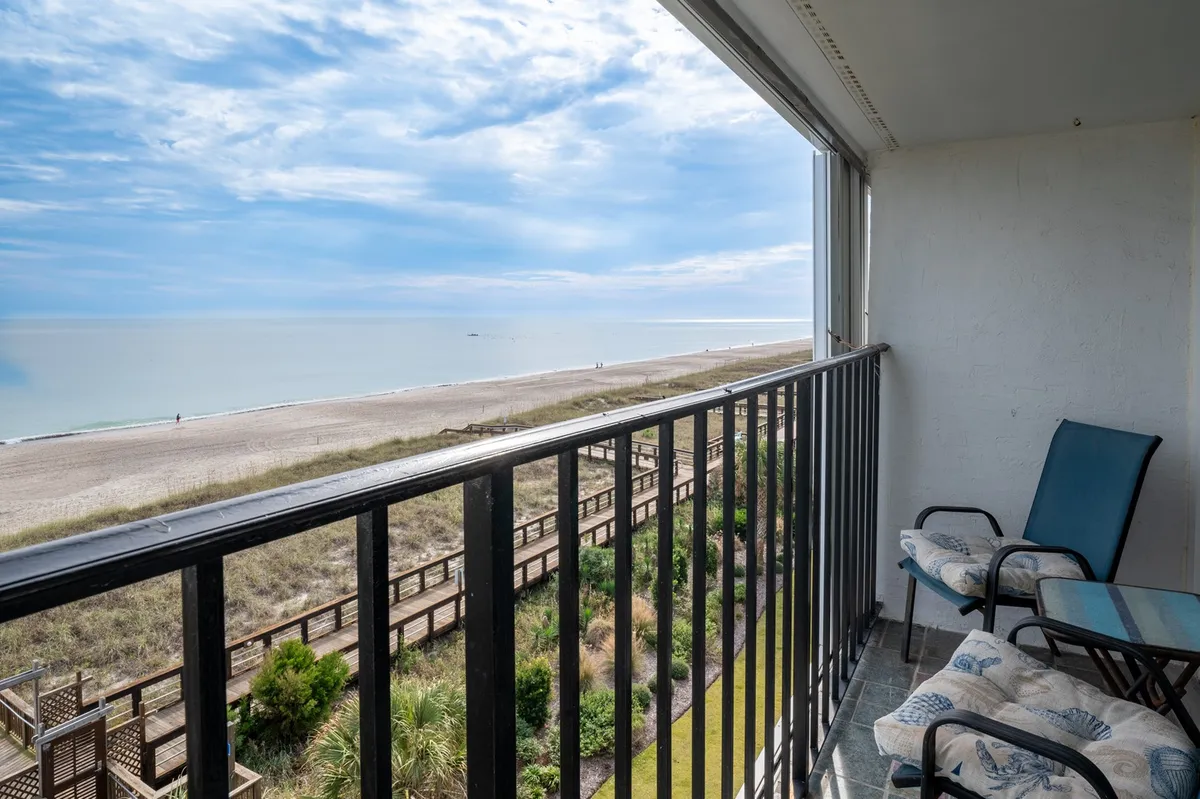 A private balcony with comfortable seating, a railing overlooking the beach, and the ocean beyond. The space is bathed in natural light, inviting moments of relaxation, reading, or sipping cocktails while waves roll in below.